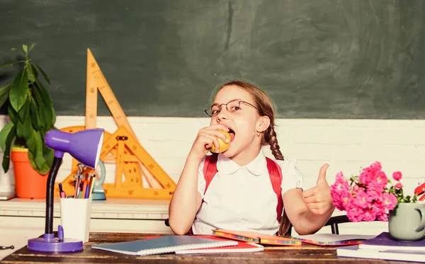 Školačka sedět stůl tabuli pozadí. Student na škole. Holčička jí jablečnou svačinku. Školní prázdniny. Klid mezi lekcemi. Vitaminová náplň. Školní životní koncept. Zdravý životní styl — Stock fotografie