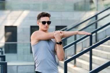 Spor salonundaki adam dışarıda egzersiz yapıyor. Erkek egzersizden önce kollarını açarak ısınır. O kasları esnek tutmak. Spor kıyafetli genç adam kolunu uzatıyor. Kaslarını hazırlıyor.