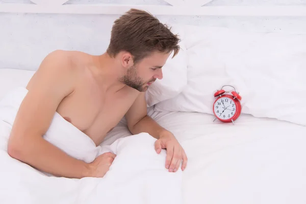 Dorminhoco. Sexy homem verificar hora na cama. Homem sem barba deitado na cama. Bonito homem acordado de manhã. Uma pessoa sonolenta. Preparar despertador. Acorde cedo. — Fotografia de Stock