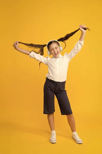 Crecer el cabello con música. Niño pequeño con peinado de cola larga y auriculares para la escuela. Niña sosteniendo peinado sobre fondo amarillo. Linda niña disfrutando de la música y su peinado. Peinado fácil — Foto de Stock