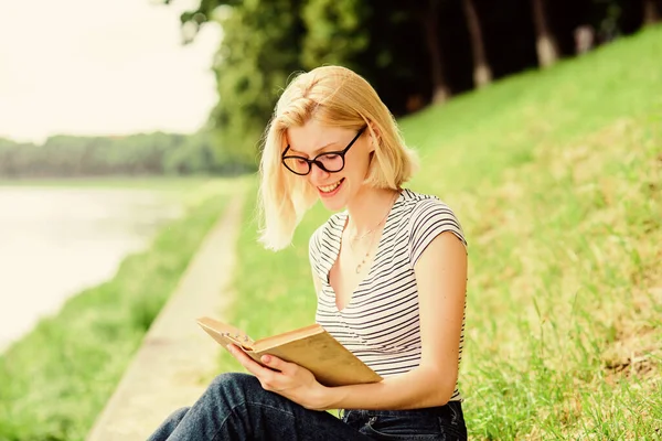 Дівчина розслабляється на березі річки після робочого дня. Жінка мріє про відпустку. Література для літніх канікул. Розслабтеся і захоплюйтеся. Літня відпустка. Дівчина сидить на зеленій траві біля річки і читає книгу — стокове фото
