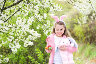 Bahçede çiçek açan ağaçlarla tavşan kulaklarıyla gülümseyen bir çocuk.