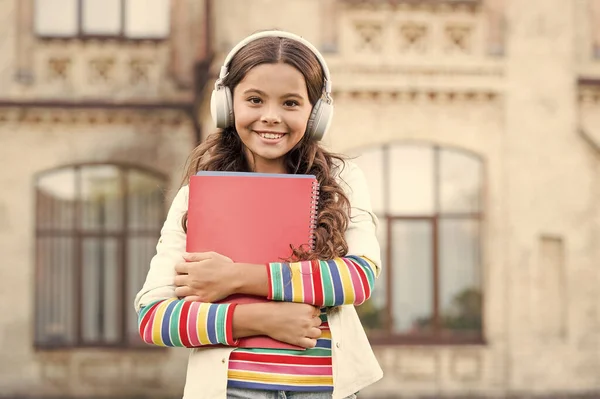 Programas de áudio para alunos. Menina feliz ouvir áudio livro tocando em fones de ouvido. Criança pequena desfrutando de aprendizagem de áudio. Criança em fones de ouvido estéreo segurando materiais de áudio para lição — Fotografia de Stock