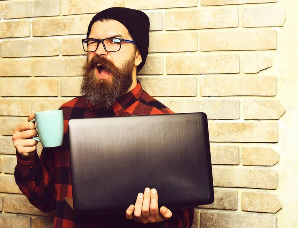 Bearded brutal caucasian hipster holding laptop with cup — Stock Photo, Image