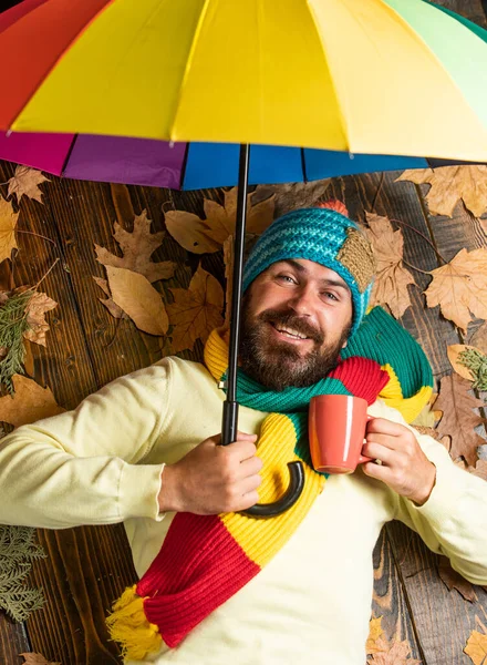 Fröhlicher Mann, der heißen Tee oder Kaffee trinkt. Herbstliche Rabatte auf Kleidung. Mann mit Regenbogen-Regenschirm. Er mag Regen. Der Wetterbericht verspricht Regen. Bärtiger Mann mit buntem Regenschirm — Stockfoto