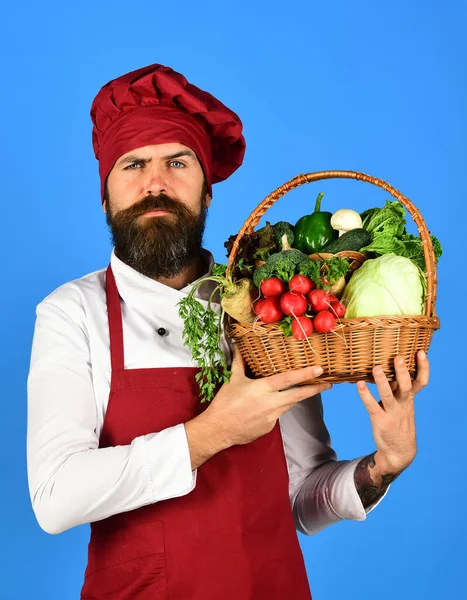 Cook with serious face in burgundy uniform holds vegetables — Stock Photo, Image