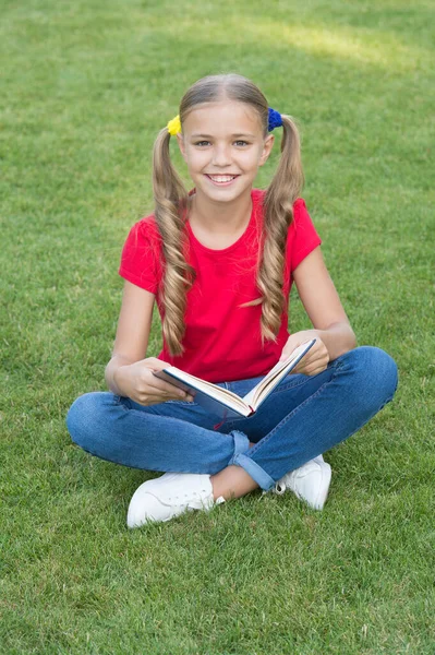 Küçük kız şirin saç stili kitap okuyor, doğa geçmişini rahatlatıyor, okula geri dönüyor. — Stok fotoğraf