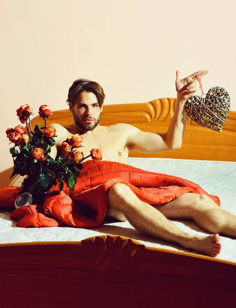 bearded macho man with roses on bed under red blanket holding bouquet