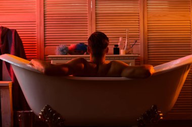 Küvette çıplak oturan güçlü omuzlu bir adam..