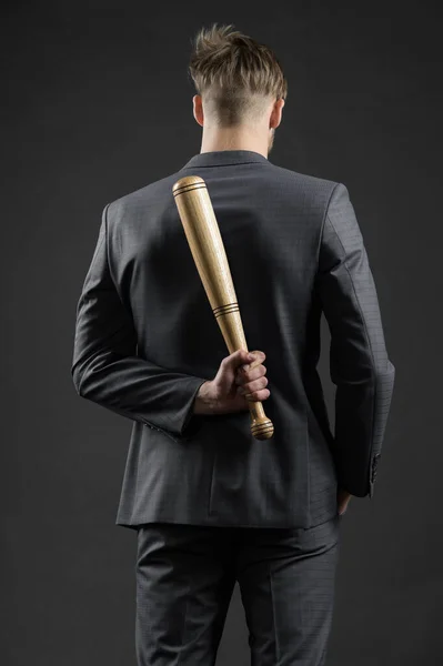 Latent aggression. Businessman or man in formal suit hides wooden bat behind back, dark background. Hidden danger concept. Man with bat hides his aggression slow down and keep calm, rear view