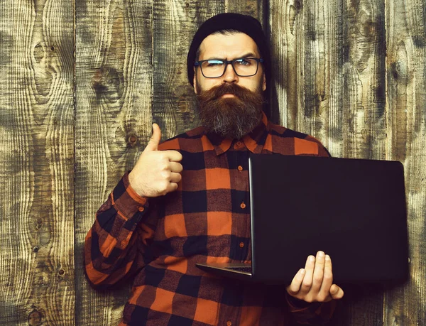 Bearded brutal caucasian hipster holding laptop on brown vintage wooden studio background — Stock Photo, Image