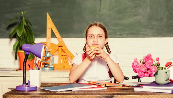 女子高生は机の黒板の背景に座る。女の子の小さな子供のアップルスナックを食べる。学校の休みだ。レッスンの合間にリラックス。ビタミン充電。健康的なライフスタイル。学校生活の概念。学校の子供の生徒 — ストック写真