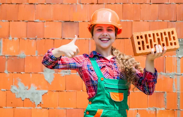 Liten arbetare i hjälm byggnad från tegel. Byggnadsbarn för arkitektur. Barn i uniform arbetar runt tegelvägg. begreppet arbetsdag. En liten flicka murare. Att lära sig bygga. tumme upp — Stockfoto