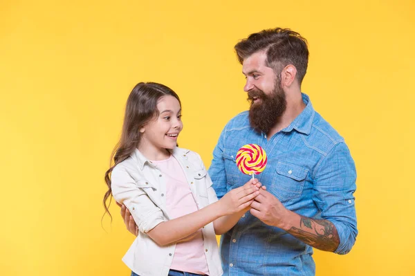 Wat een verrassing. Een man met baard geeft lolly aan een verrast kind. Zoetekauw familie gele achtergrond. Sweetshop en snoepgoed. Snoepwinkel. Zoet dat spreekt. — Stockfoto
