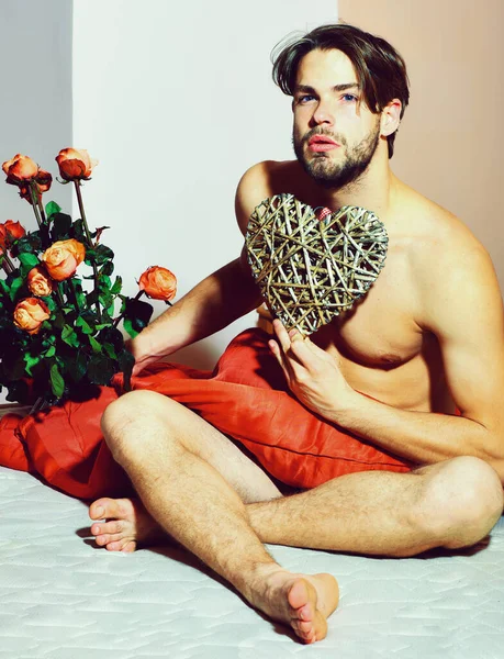 Macho barbudo con rosas en la cama debajo de una manta roja con ramo de rosas naranjas y corazón decorativo de San Valentín — Foto de Stock