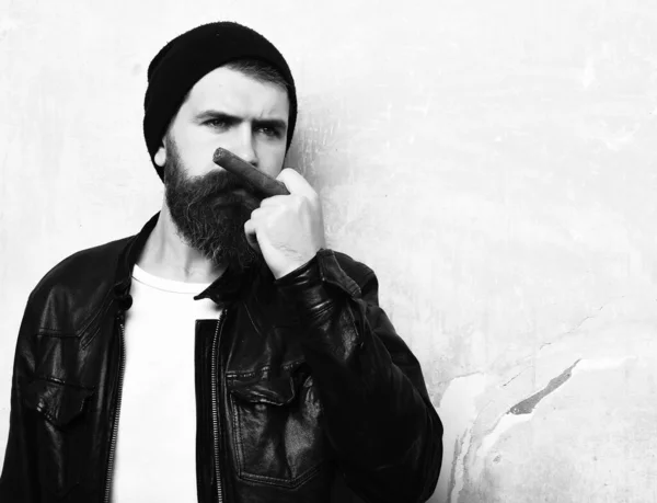 Bearded brutal caucasian hipster smoking cigar on battered wall background, copy space
