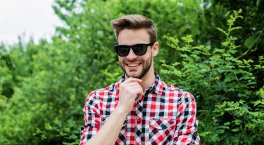 Tarzına güveniyor. Gülümseyen adam kareli gömlek ve güneş gözlüğü takıyor. Yaz tatili konsepti. Açık havada kıllı yakışıklı bir adam. Erkek modası ve güzellik. Seksi sakallı maço. günlük kıyafet.