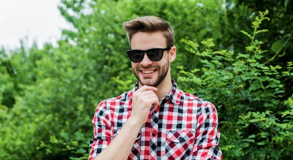 Tarzına güveniyor. Gülümseyen adam kareli gömlek ve güneş gözlüğü takıyor. Yaz tatili konsepti. Açık havada kıllı yakışıklı bir adam. Erkek modası ve güzellik. Seksi sakallı maço. günlük kıyafet.