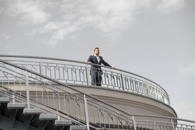 Kendine güvenen işadamı bulutlu mavi gökyüzünün balkonunda duruyor. Resmi kıyafetli adam açık havada giyiniyor. Geleceğe bakıyorum. Yeni olasılıkları düşünüyorum. Özgüven ve karizma. Erkek görünüşü. Gelmiş geçmiş en iyi yönetici.
