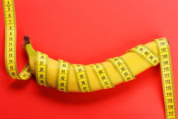 Banana with tape for measuring figure. Yellow tape around banana isolated on red background. — Stock Photo, Image