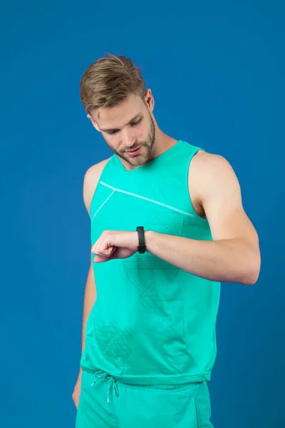 El deportista mira el reloj de fitness sobre fondo violeta. Hombre con reloj inteligente deportivo en ropa deportiva azul. Seguimiento de la actividad y el entrenamiento. Nueva tecnología para un estilo de vida saludable. Deporte o fitness y bienestar — Foto de Stock