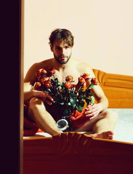 Macho barbudo con rosas en la cama bajo manta roja con ramo de rosas naranjas en jarrón transparente — Foto de Stock