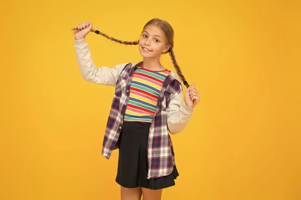 Longo e trançado. Sorriso de criança adorável com longas tranças de cabelo loiro. Menina pequena feliz segurar longo trançado penteado fundo amarelo. Estimulante de crescimento de cabelo longo — Fotografia de Stock