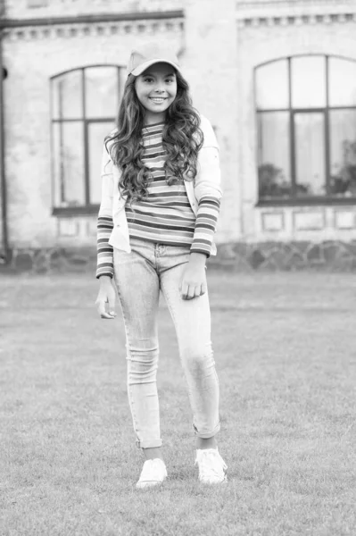 Menina feliz com cabelo encaracolado usando boné. Criança sorrindo ao ar livre. Beleza e moda. Caminhando gramado verde. Costumava andar por aí. A estudante usa boné. Adolescente em estilo casual e boné. Felicidade infantil — Fotografia de Stock