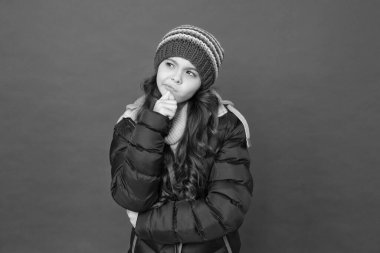 Kış rüyası. Sıcak kış kıyafetleri içinde düşünceli bir çocuk. Çocuklar için mevsimlik moda. Küçük güzel kırmızı arka plan. Küçük kız kabarık ceket ve örgü şapka. Soğuk havada kendine dikkat et.