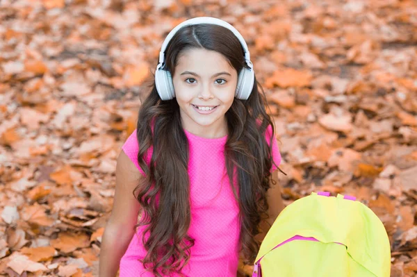 幸せな子供時代。子供は秋の公園で音楽を聴く。秋は勉強の時間です。学校に戻って。オンライン教育の概念です。子供は紅葉の中に座っている。学校のバッグを持ったイヤホンの女の子 — ストック写真