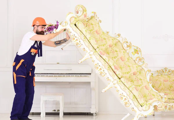 Delivery service concept. Loader moves sofa, couch. Man with beard, worker in overalls and helmet lifts up sofa, white background. Courier delivers furniture in case of move out, relocation.