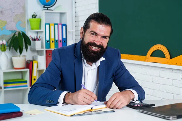 Happy školní učitel s notebookem dívá do kamery a sedí ve třídě, školní vzdělávání — Stock fotografie