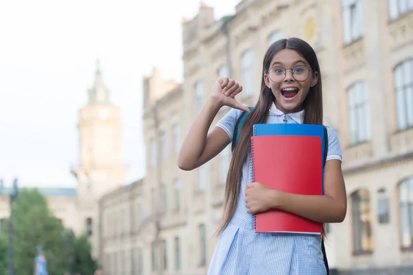 Szczęśliwe małe dziecko okulary girlin wskazując palcem kciuka w książkach szkolnych na zewnątrz, informacje, przestrzeń kopiowania — Zdjęcie stockowe