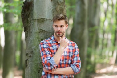 Güven. Tüylü adam kareli gömlek giyiyor. Sıradan moda tarzı. Ormandaki kaslı adam. Açık havada düğmesiz gömlekli seksi maço. Genç traşsız adam saçını taramış. Erkek güzelliği ve moda