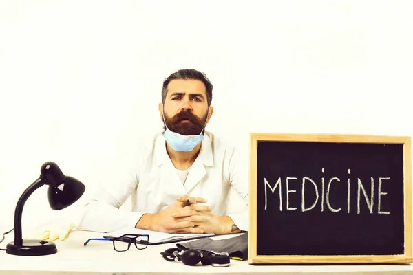 Biały lekarz z brodą siedzący przy stole z napisem "Medycyna" — Zdjęcie stockowe