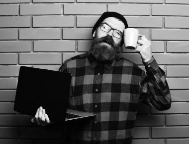 Sakallı, beyaz tenli hippi elinde bardakla dizüstü bilgisayar tutuyor.