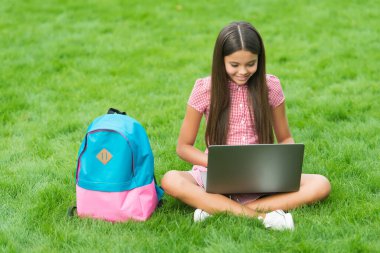 Bilgisayarda çalışan mutlu çocuk. Online eğitim. Okula dönüyorum. Genç kız parktaki yeşil çimlerde bilgisayar kullanıyor. Sırt çantası ve defteri olan bir çocuk. Modern hayatta yeni teknoloji. çalışma