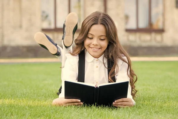 Grundbildung. Liebenswertes kleines Mädchen lernt lesen. Schuluniform für Schulmädchen auf dem Rasen mit Lieblingsbuch. Studienkonzept. Extraschulische Lektüre. Niedliches kleines Kind liest Buch im Freien — Stockfoto