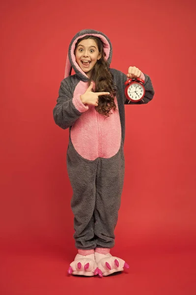 计算时间。现在是周末可爱的兔子拿着闹钟.穿着兔子服装的小女孩。兔子kigurumi的孩子。快乐的女孩兔子睡衣。休息和放松。兔子小孩的红色背景。小动物 — 图库照片