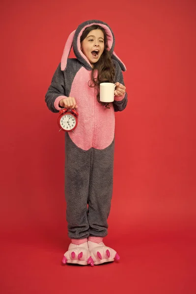 Drinking coffee energy. Morning routines. Adorable bunny hold alarm clock. Small girl in bunny costume. Child rabbit kigurumi. Girl bunny pajamas. Bunny kid red background. Baby animal. Counting time