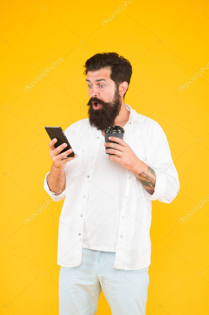 guy with beard and moustache hold mobile phone. communication. morning routine. surprised bearded man has incoming phone call while drink coffee. brutal hipster drinking takeaway beverage from cup