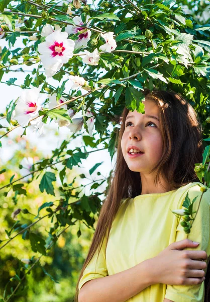 Estilo de moda de primavera de criança. infância feliz. criança bonita alegre na árvore florescente ao ar livre. férias. moda de verão e beleza. bela menina adolescente com hibisco arbusto flor — Fotografia de Stock