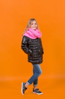 Uzun sarı saçlı, siyah ceketli, atkılı, kotlu, spor ayakkabılı çocuk.