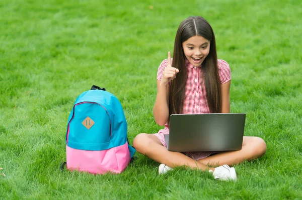 Geïnspireerd gelukkig kind met behulp van computer voor online schoolstudie zitten in park op groen gras met rugzak, onderwijs — Stockfoto