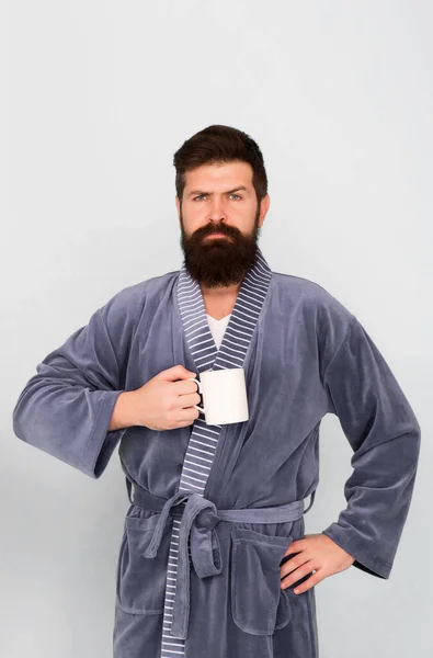 Bearded man in bathrobe with mug. Breakfast concept. Man with beard in blue bathrobe enjoy morning coffee or tea. Guy in bath clothes hold tea coffee. Start new day. Every morning begins with coffee
