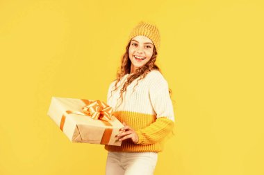 Mutlu Noeller ve mutlu tatiller. Mutlu kış tatilleri. Neşeli küçük kız örgü şapka ve kazak örüyor. Çocuk hediye kutusunu sarı arka planda tutuyor. Xmas hediye alışverişi. Noel 'in aydınlık olsun.