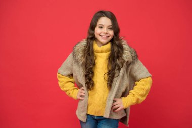 Gülümseyen genç kız uzun kıvırcık saçlı kalın örgü süveter ve kırmızı arka planda kürk yelek giyiyor, sonbahar modası.