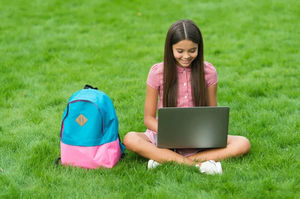 Glad unge med bärbar dator för online skola utbildning sitter i parken på grönt gräs med ryggsäck, barndom — Stockfoto