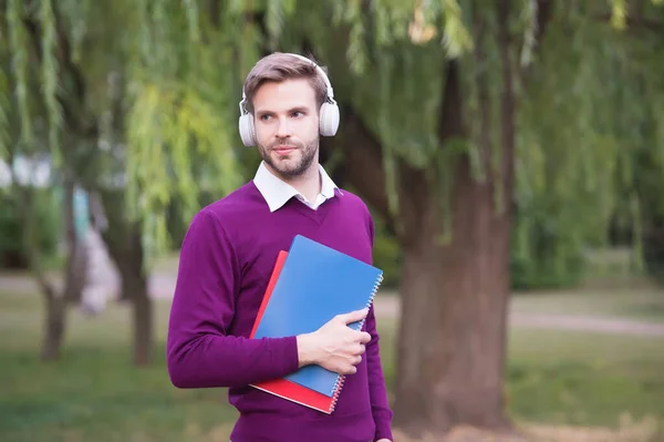 Étudiant obtenir des informations du livre. concept d'apprentissage en ligne. homme écouter de la musique dans les écouteurs et tenir un cahier. beau gars dans les écouteurs se détendre dans le parc. technologie moderne d'éducation à la vie — Photo