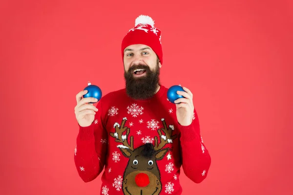 Concepto de año nuevo. Fiesta de Navidad. Traje de invierno. Buenas vibraciones. Decoraciones de invierno. Actividades festivas divertidas. Hipster sonriente hombre barbudo alegre usar suéter de invierno y sombrero sostienen bolas de fondo rojo — Foto de Stock